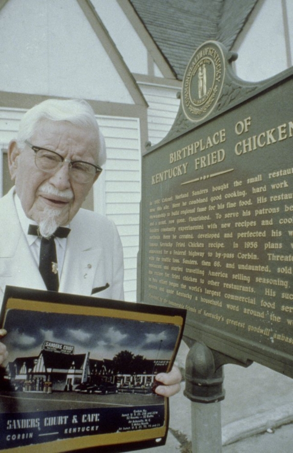 ภาพถ่ายปี 1970 ผู้พันแซนเดอร์สโชว์รูปภาพ Sanders Café ข้างๆ แผ่นป้าย “Birthplace of Kentucky Fried Chicken” 